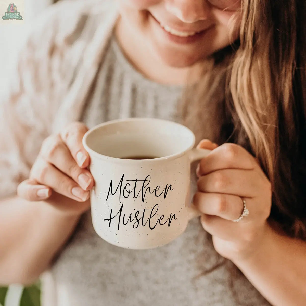 A woman with long brown hair smiles warmly, holding the Mother Hustler 14.5oz mug. Her ring and cozy vibe show she’s a true mom boss, fueled and motivated by her favorite coffee mug.