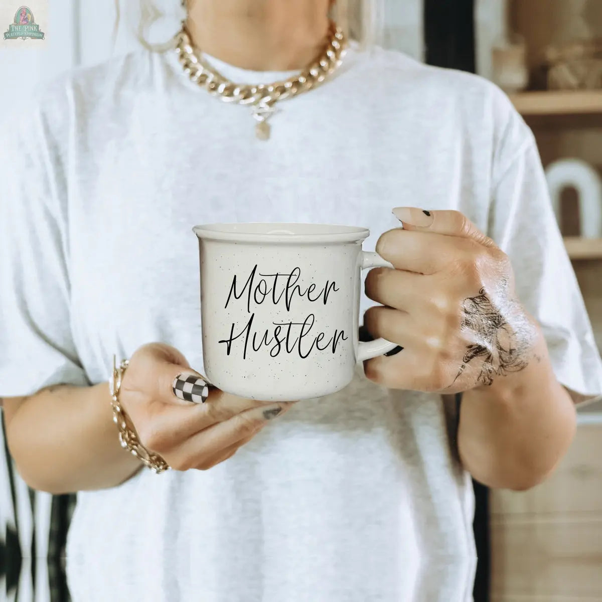A person in a white shirt with gold jewelry holds the Mother Hustler 14.5oz mug, featuring handwritten text. One hand shows black ink marks and checkered nails, all set against a softly blurred indoor background.