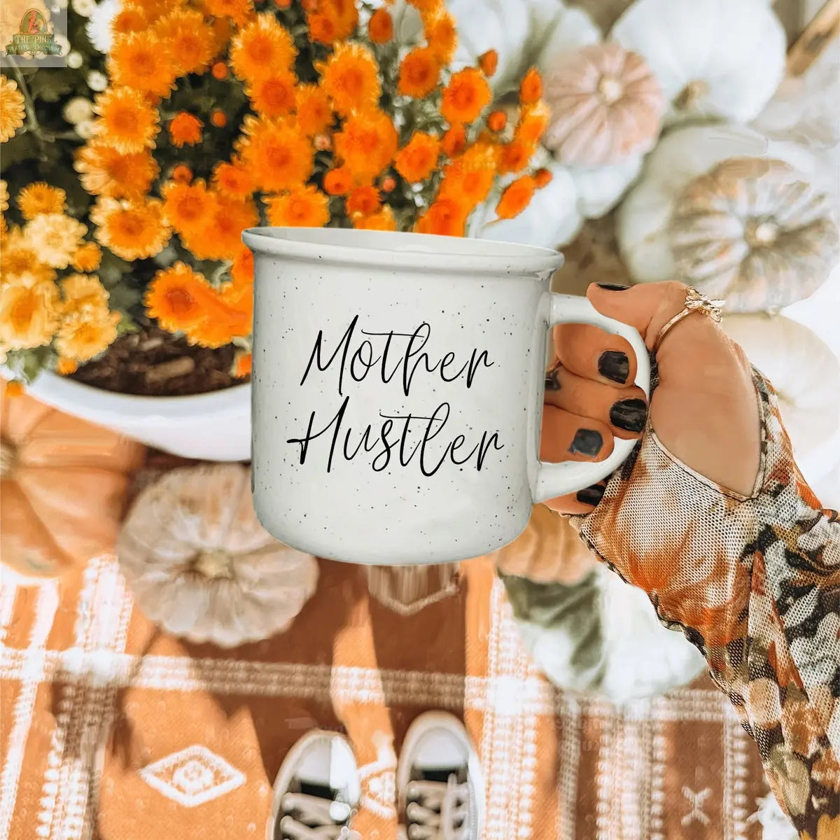 Someone holds a "Mother Hustler 14.5oz" motivational coffee mug filled with flowers, surrounded by pumpkins.
