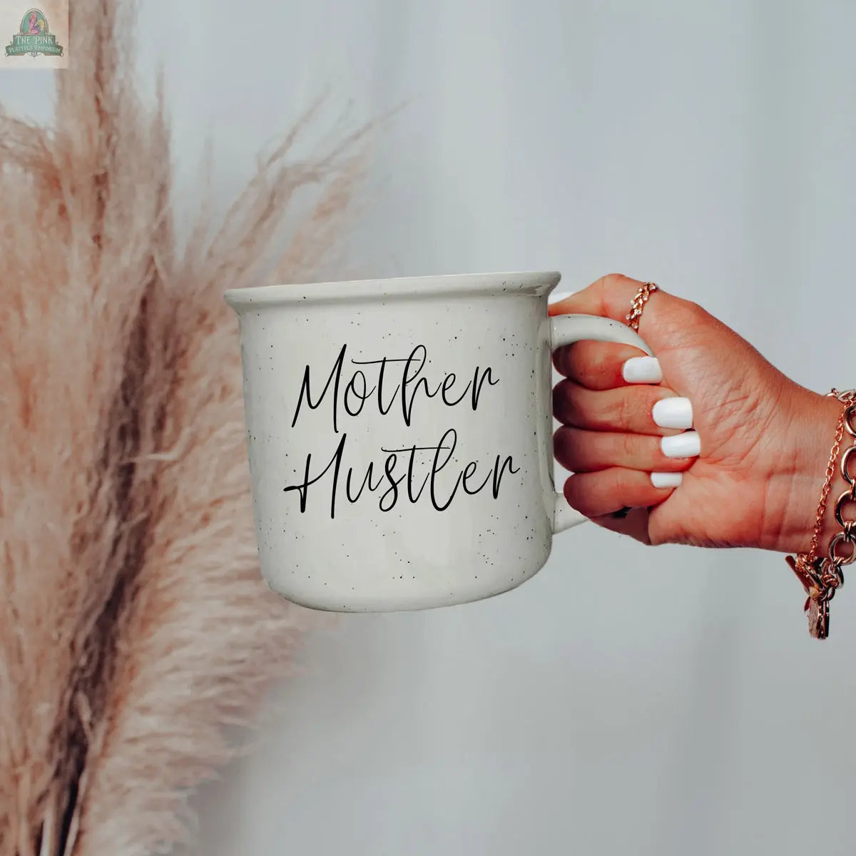 A hand with white nail polish and gold rings holds the Mother Hustler 14.5oz mug. Beige pampas grass in the background adds a chic touch to this inspiring mom boss moment.