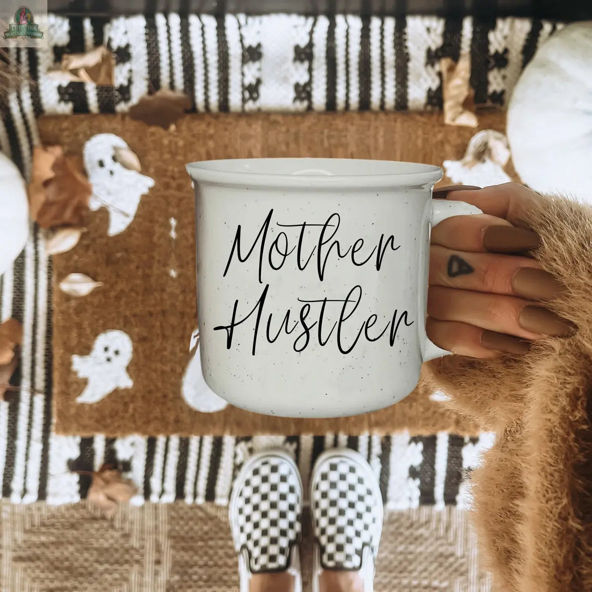 A hand with manicured nails holds the Mother Hustler 14.5oz mug above a fall-themed ghost doormat. The person, wearing checkered slip-ons, stands on a patterned rug scattered with autumn leaves.