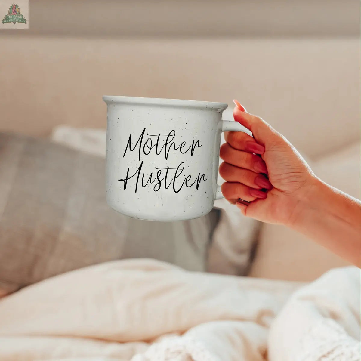 A hand holds the Mother Hustler 14.5oz mug, featuring bold black script on white ceramic. Set on a bed with beige and gray pillows, this mug brings motivational style to any cozy space.