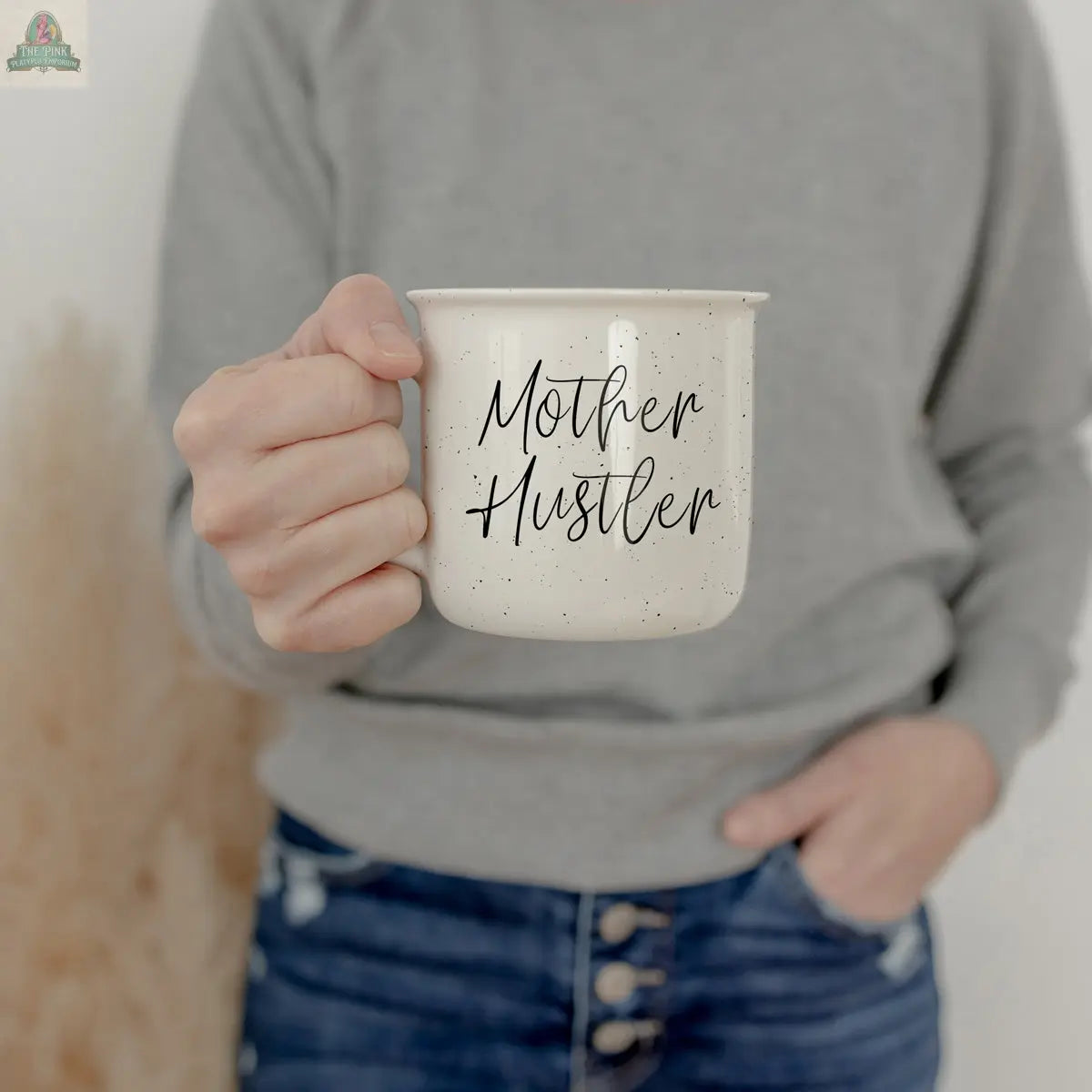 A person in a gray sweater and blue jeans holds the Mother Hustler 14.5oz mug, featuring motivational black text that reads “Mother Hustler.”.