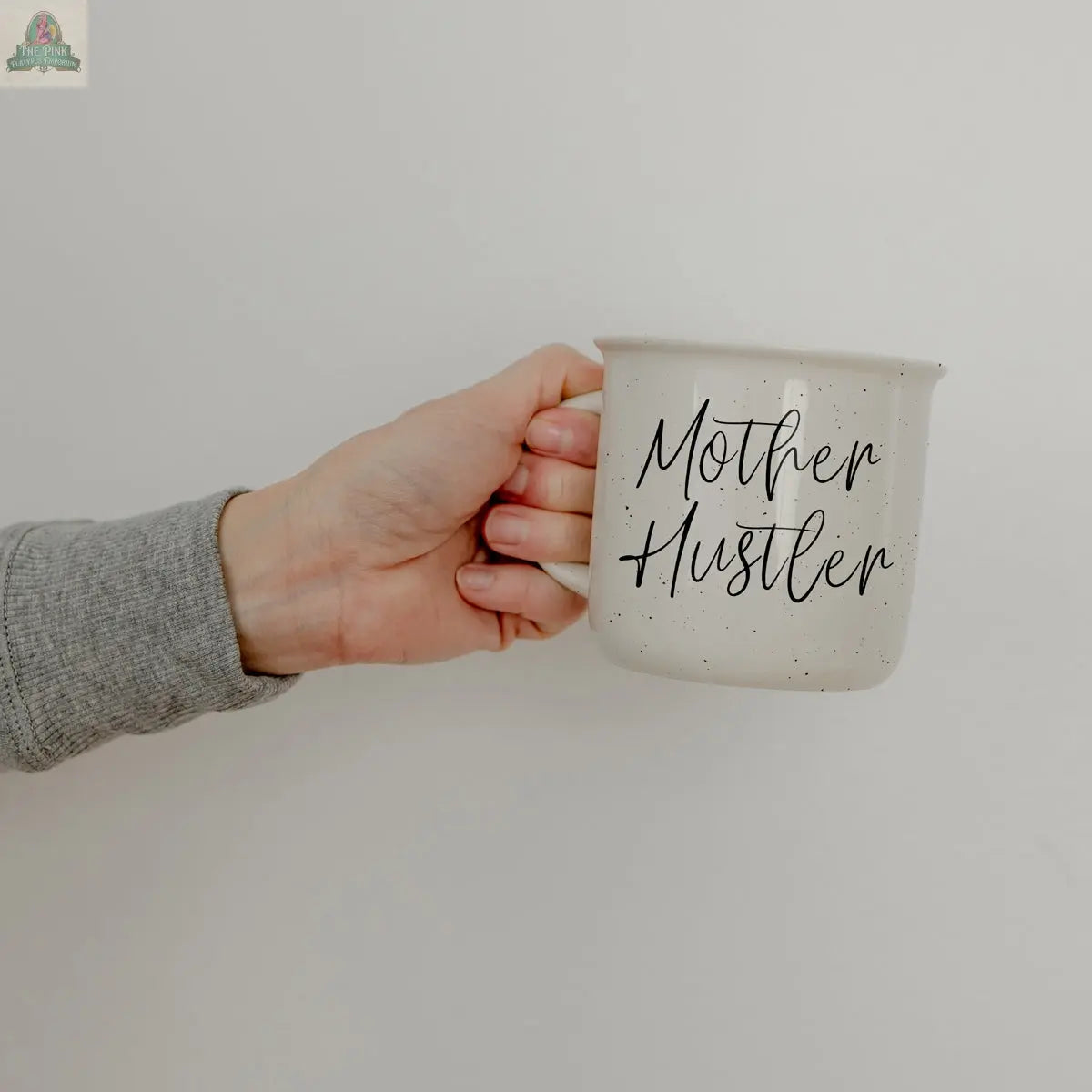 A person in a gray sleeve holds the Mother Hustler 14.5oz white coffee mug, featuring black speckles and “Mother Hustler” in cursive. The plain, light wall serves as the background.