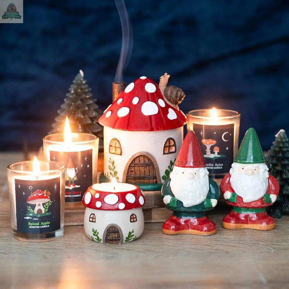 A whimsical arrangement features a Mushroom House Resin Tealight Holder, gnome figurines, two small trees, and festive jar candles—perfect winter forest home decor set against a dark blue background.
