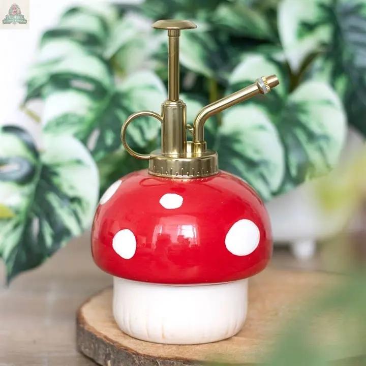 The Mushroom Shaped Plant Mister, featuring a red and white spotted ceramic base and brass nozzle, sits on a wooden surface among green leafy plants, bringing whimsical cottagecore charm to an enchanted forest setting.