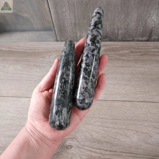 A hand holds two polished Keystone Crystals Mystic Merlinite Round Massage Wands, showing gray and black patterns against a light wooden background.