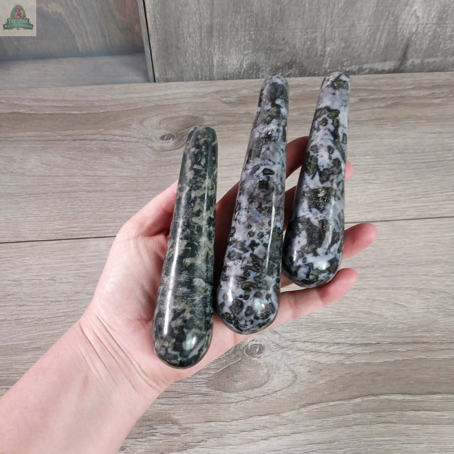 A hand holds three Keystone Crystals Mystic Merlinite Round Massage Wands, displaying their speckled black, gray, and white pattern against a wooden background.