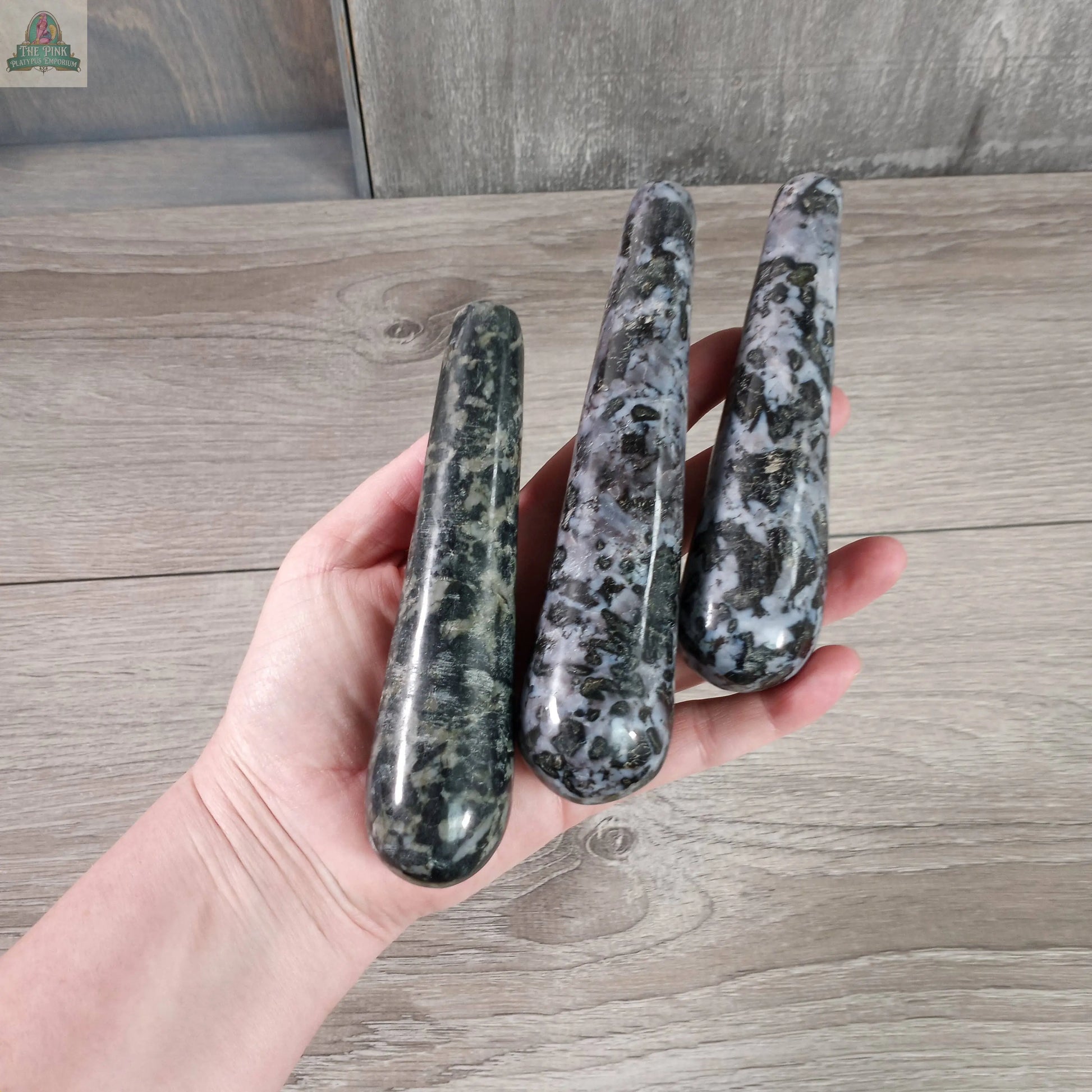 A hand holds three Keystone Crystals Mystic Merlinite Round Massage Wands, displaying their speckled black, gray, and white pattern against a wooden background.
