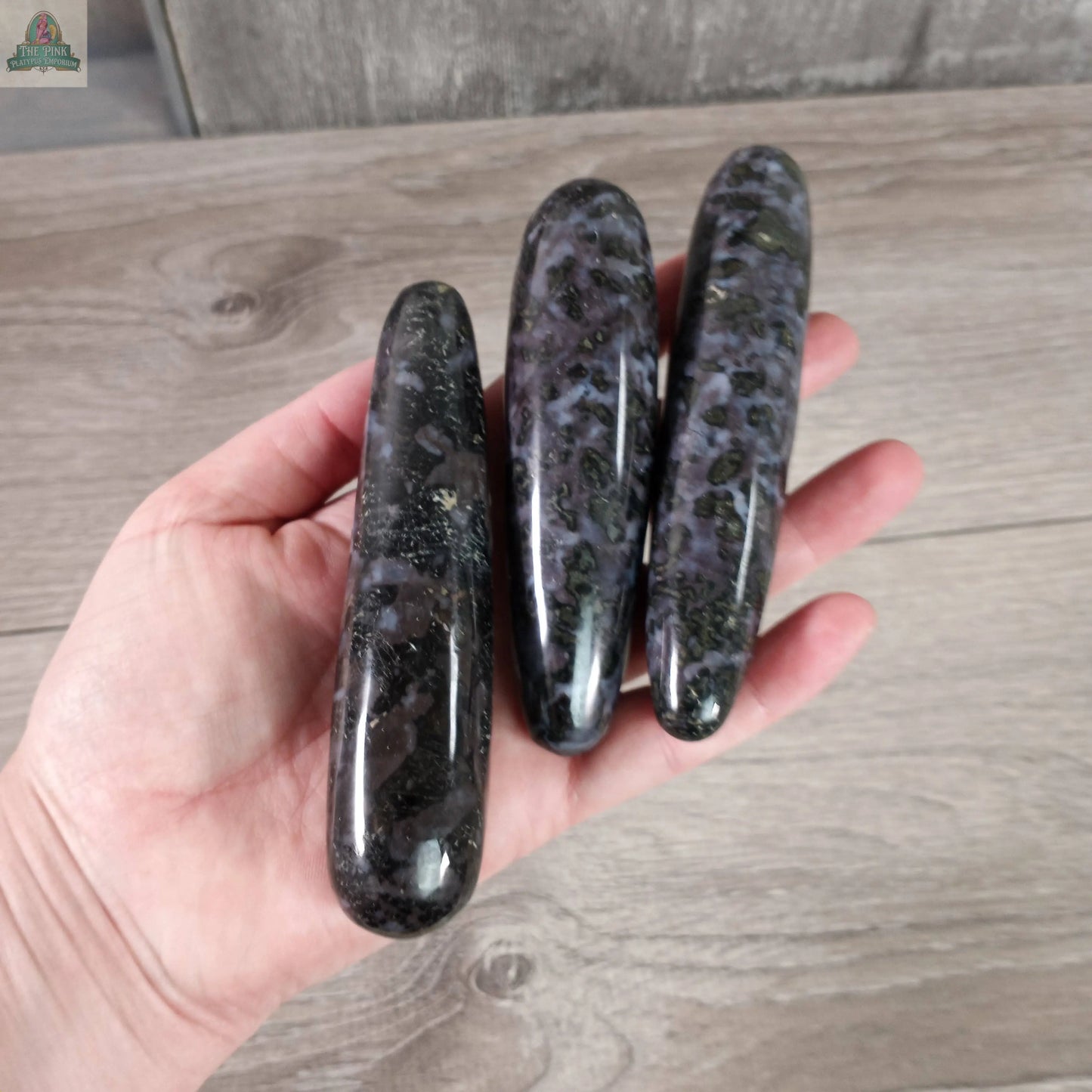 A hand holds three polished, round Mystic Merlinite Massage Wands by Keystone Crystals, featuring mottled purple and black patterns, displayed against a light wooden background.