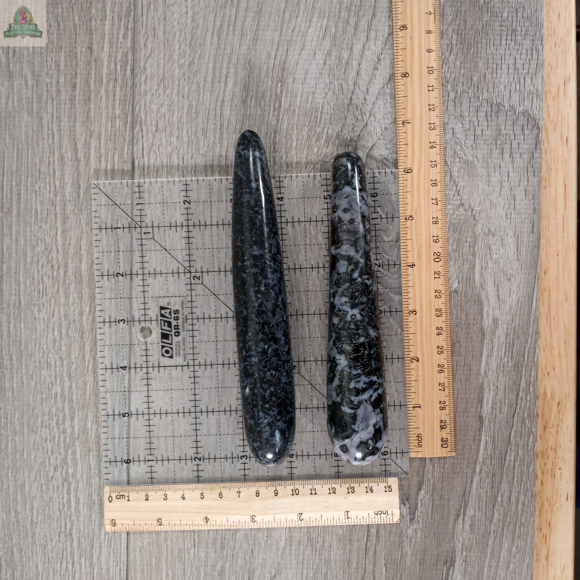 Two polished Mystic Merlinite Round Massage Wands by Keystone Crystals, in black-and-gray, rest on wood beside a clear square ruler and two wooden rulers for scale. The wands are shaped like elongated ovals.