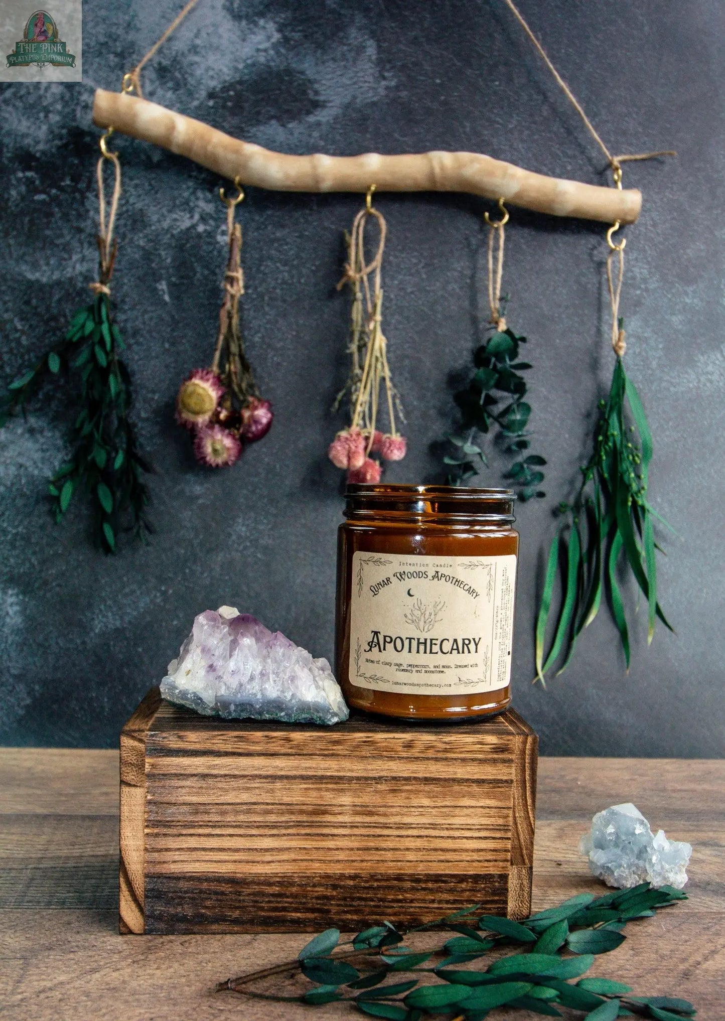 The NEW Apothecary jar rests on a wooden box next to two crystals and a spellbook candle, while dried herbs and flowers hang on a stick in the background against a textured gray wall.