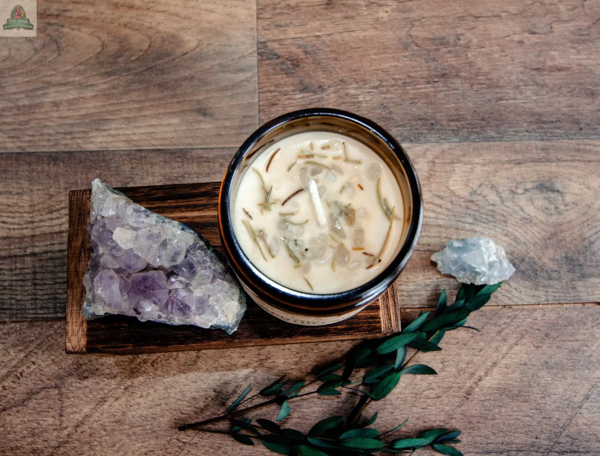 The NEW Apothecary herbal candle in a glass jar rests on a wooden tray beside a large amethyst, a small clear crystal, and greenery, creating a tranquil metaphysical display on a wooden surface.