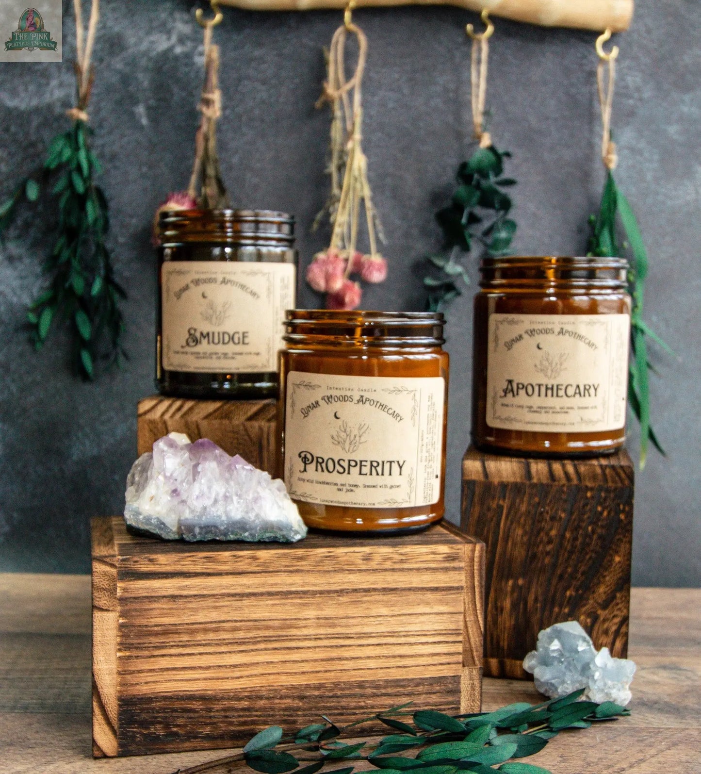 The amber glass candle “NEW Prosperity” sits on a wooden block, surrounded by crystals, greenery, and a jade charm. Dried herbs hang in the background, enhancing this tranquil display.
