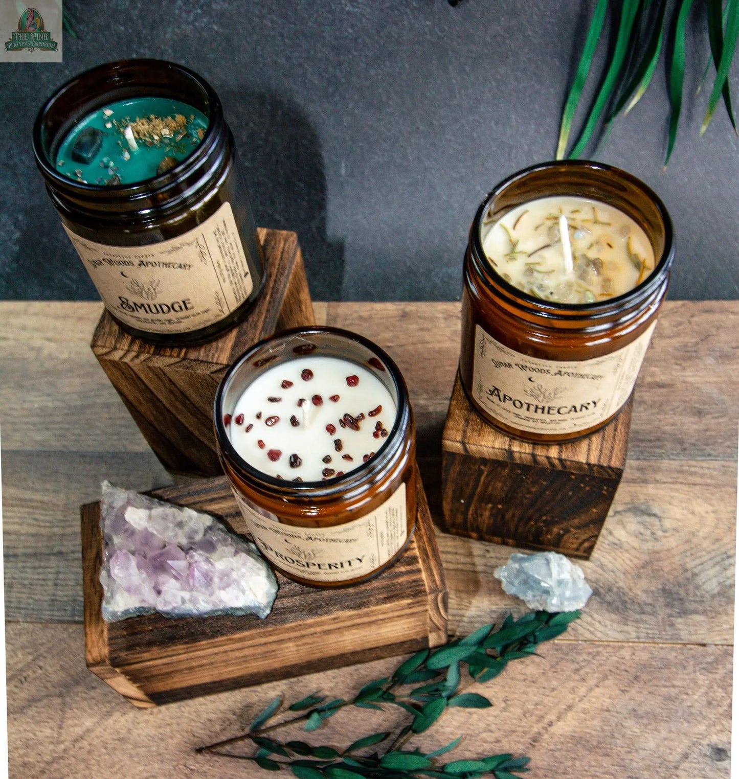 The NEW Prosperity brown glass candle is displayed on wooden blocks with decorative stones, green leaves, and a jade prosperity charm, set on a wooden surface against a dark background.