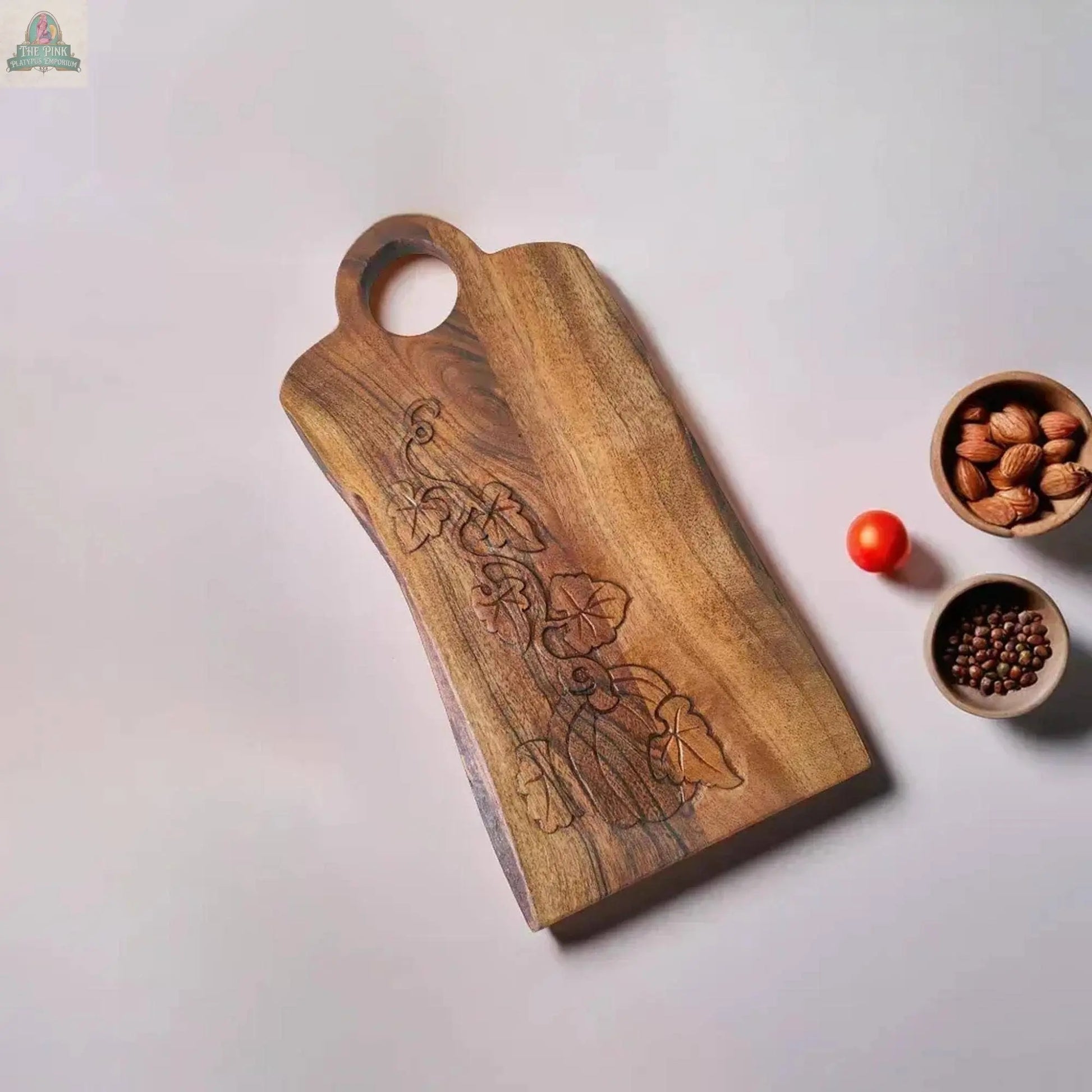 A Young's Natural Wood Charcuterie Board with a carved pumpkin design rests on a light surface beside a red cherry tomato, a small bowl of almonds, and a small bowl of black peppercorns.