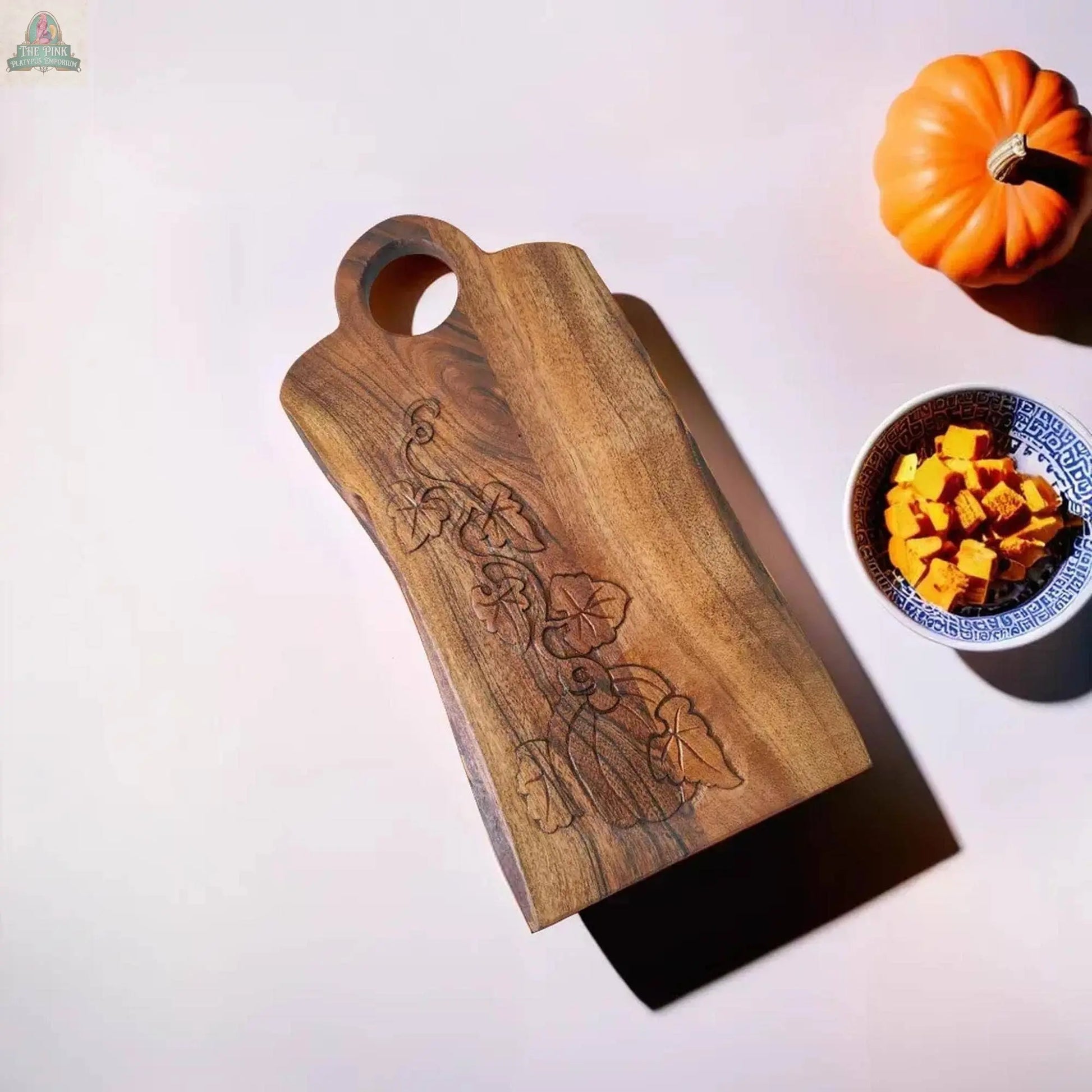 Young's Natural Wood Charcuterie Board With Carved Pumpkin Design rests on a light surface, accompanied by a small orange pumpkin and a blue-patterned bowl filled with diced pumpkin pieces.