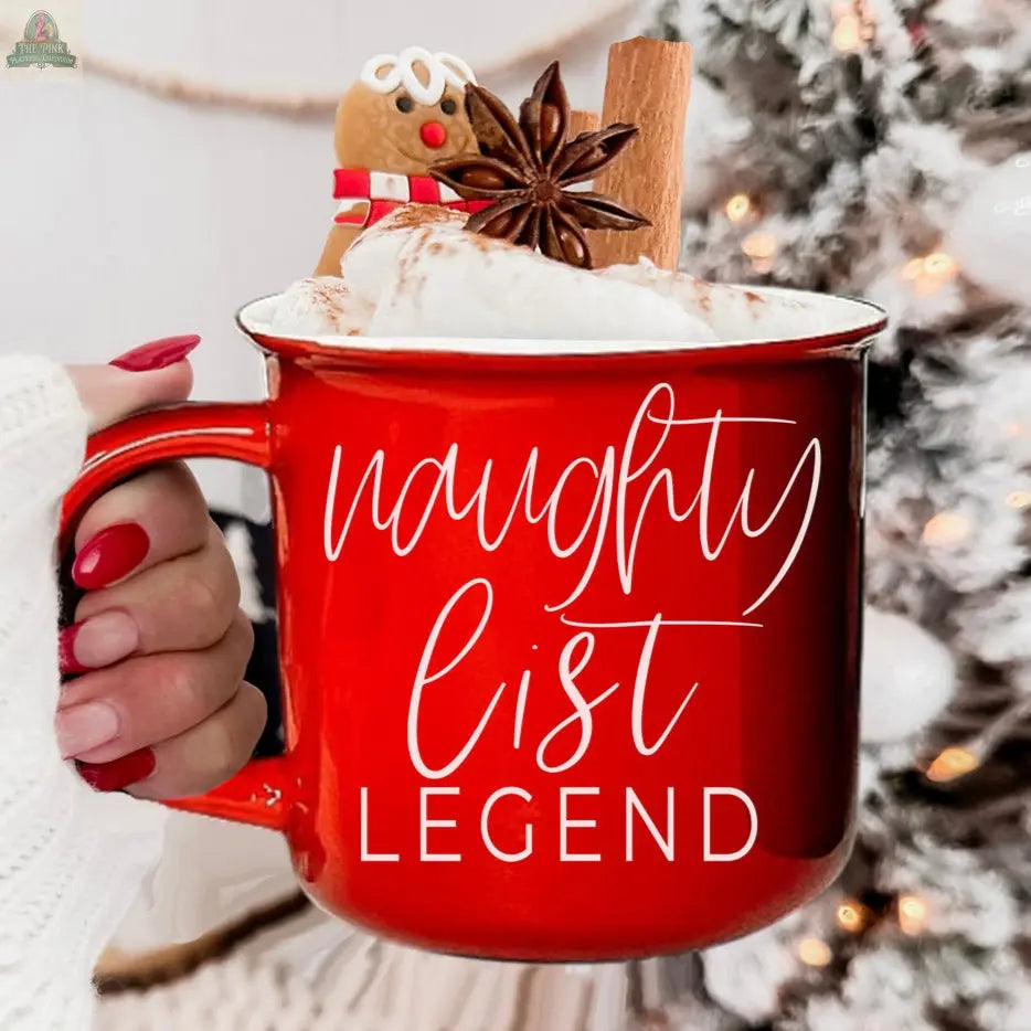 A hand with red nail polish holds a red Naughty Legend Mug filled with whipped cream, a cinnamon stick, star anise, and a gingerbread cookie. A blurred, decorated Christmas tree is visible in the background.