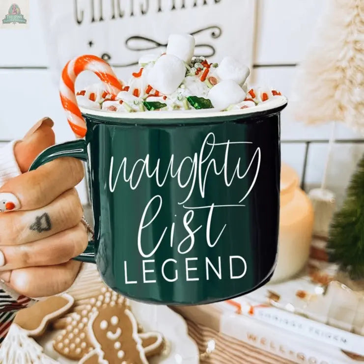A hand with festive nail art holds a Naughty Legend Mug filled with hot chocolate, whipped cream, marshmallows, sprinkles, and a candy cane. Holiday cookies and decorations create a cozy backdrop for this cheerful holiday treat.