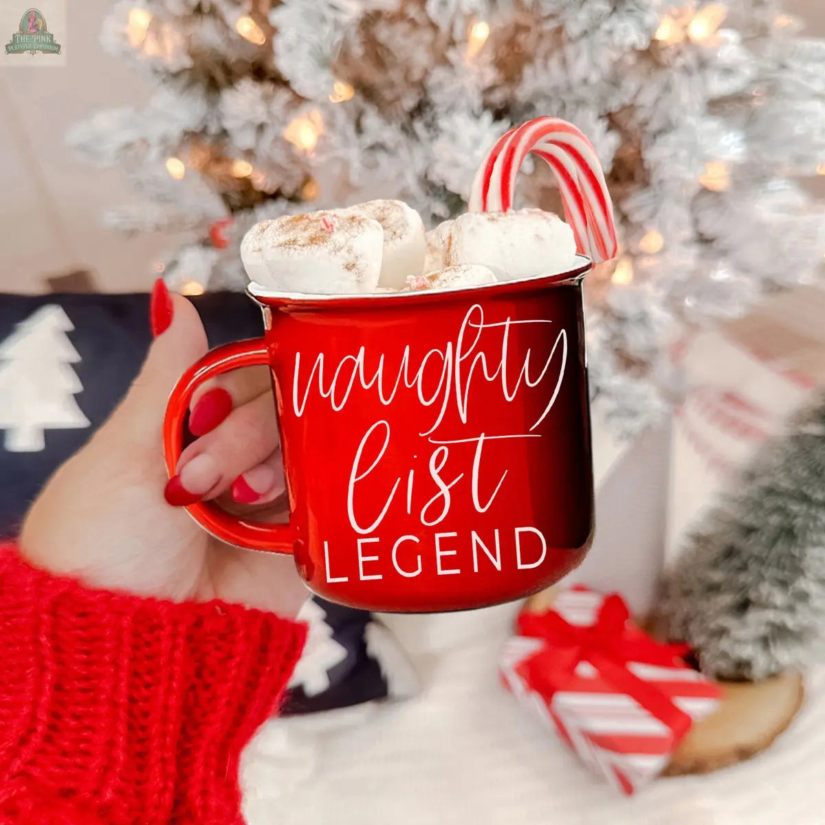 A hand with red nails holds the Naughty Legend Mug filled with hot chocolate, marshmallows, and a candy cane. “Naughty list legend.” is printed on the mug. A snowy Christmas tree and wrapped gift appear in the background.