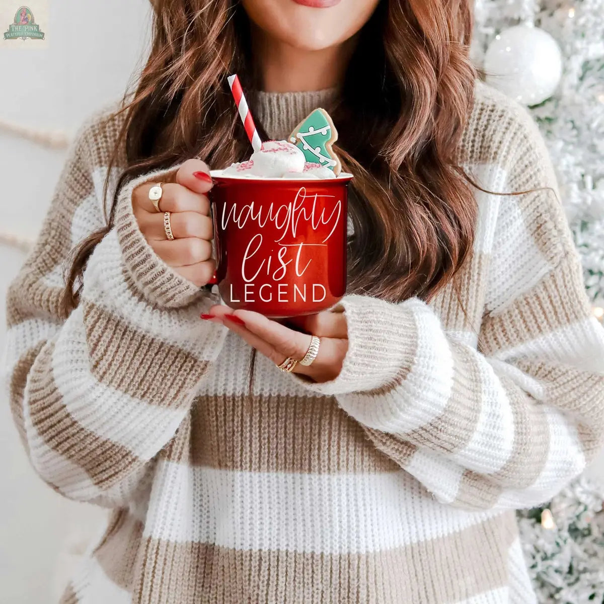 A woman in a beige and white striped sweater holds a Naughty Legend Mug topped with marshmallows, a candy cane, and a gingerbread cookie, standing in front of a decorated Christmas tree.