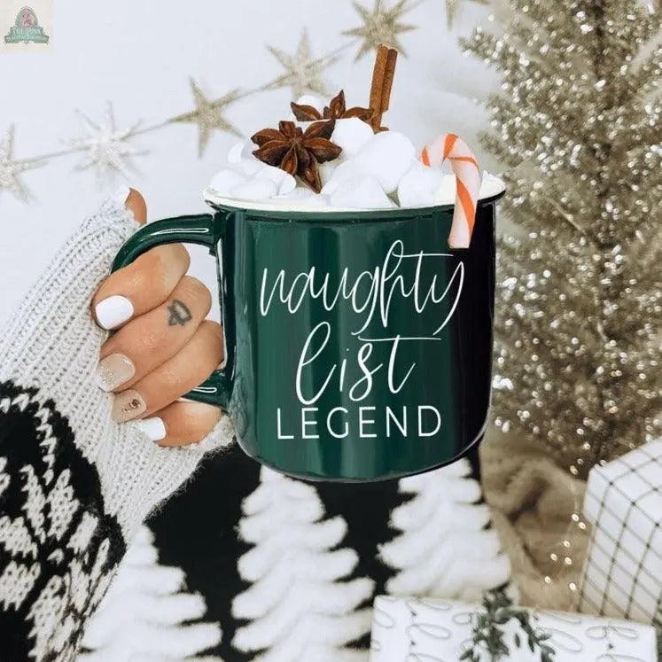 A hand with festive nail art holds a ceramic Naughty Legend Mug filled with hot chocolate, topped with marshmallows, a candy cane, a cinnamon stick, and star anise. Warm holiday decor glows softly in the background.