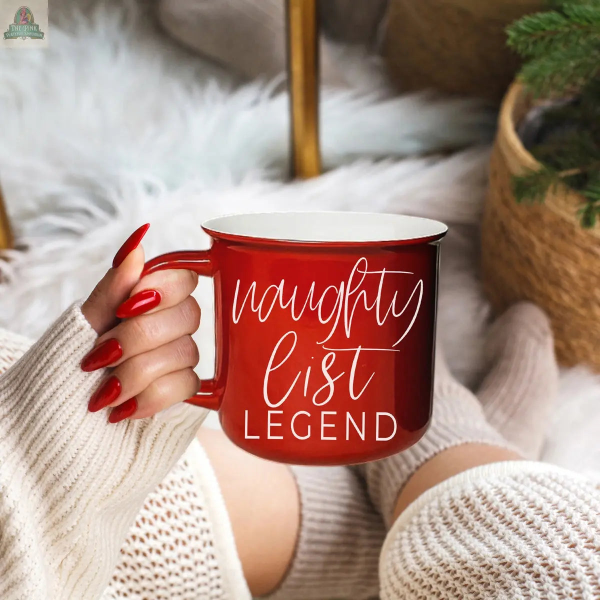 A person with red painted nails holds the Naughty Legend Mug while cozy in a white sweater and socks, sitting near a fluffy rug and a basket with greenery.