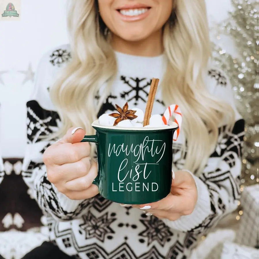 A smiling woman in a black and white sweater holds the Naughty Legend Mug, a green ceramic campfire mug with “naughty list LEGEND” in white script, filled with hot chocolate, marshmallows, a candy cane, cinnamon stick, and star anise.