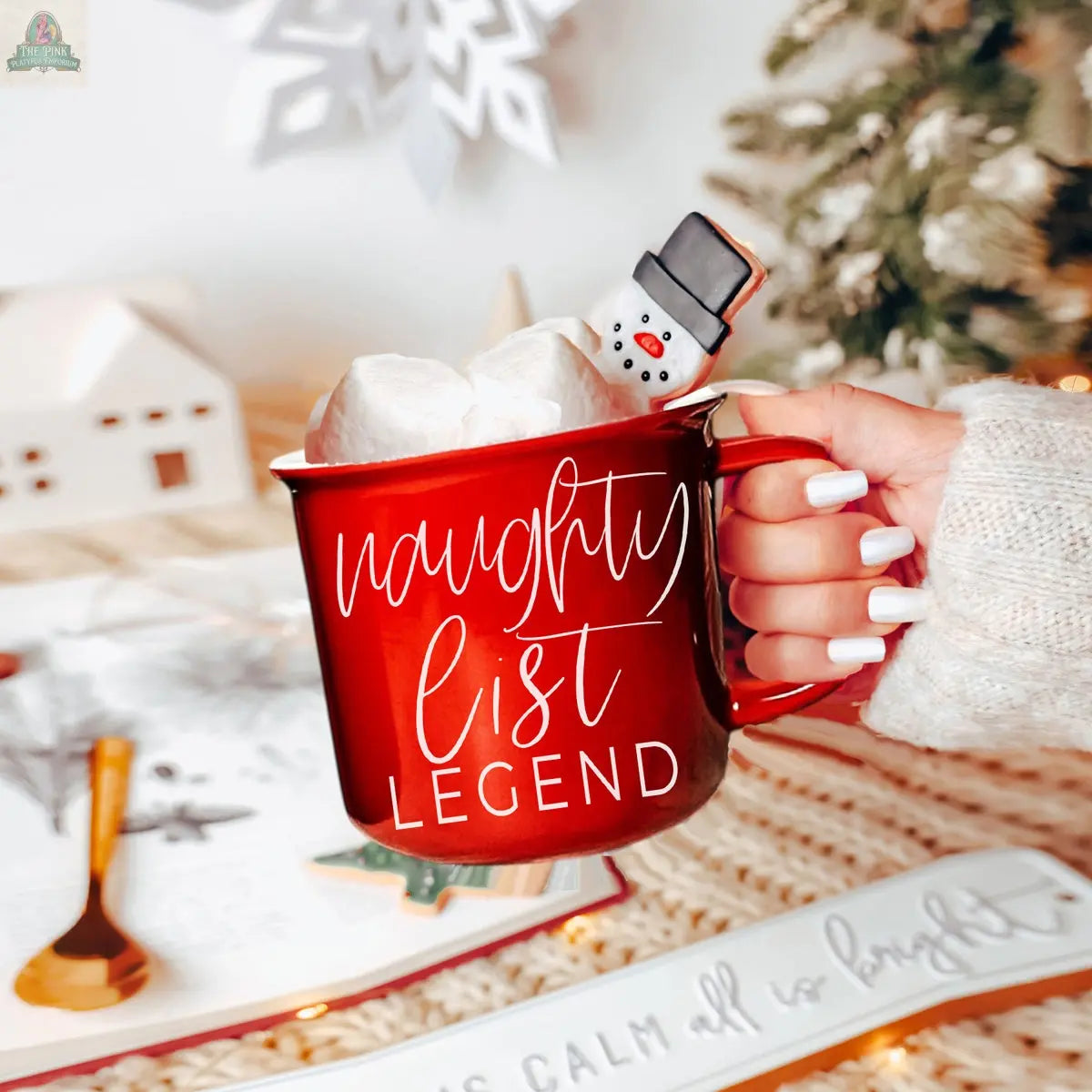 A hand with white-painted nails holds a red Naughty Legend Mug filled with marshmallows and a snowman treat, surrounded by cozy holiday decor, a Christmas tree, and a snowy village for a festive touch.