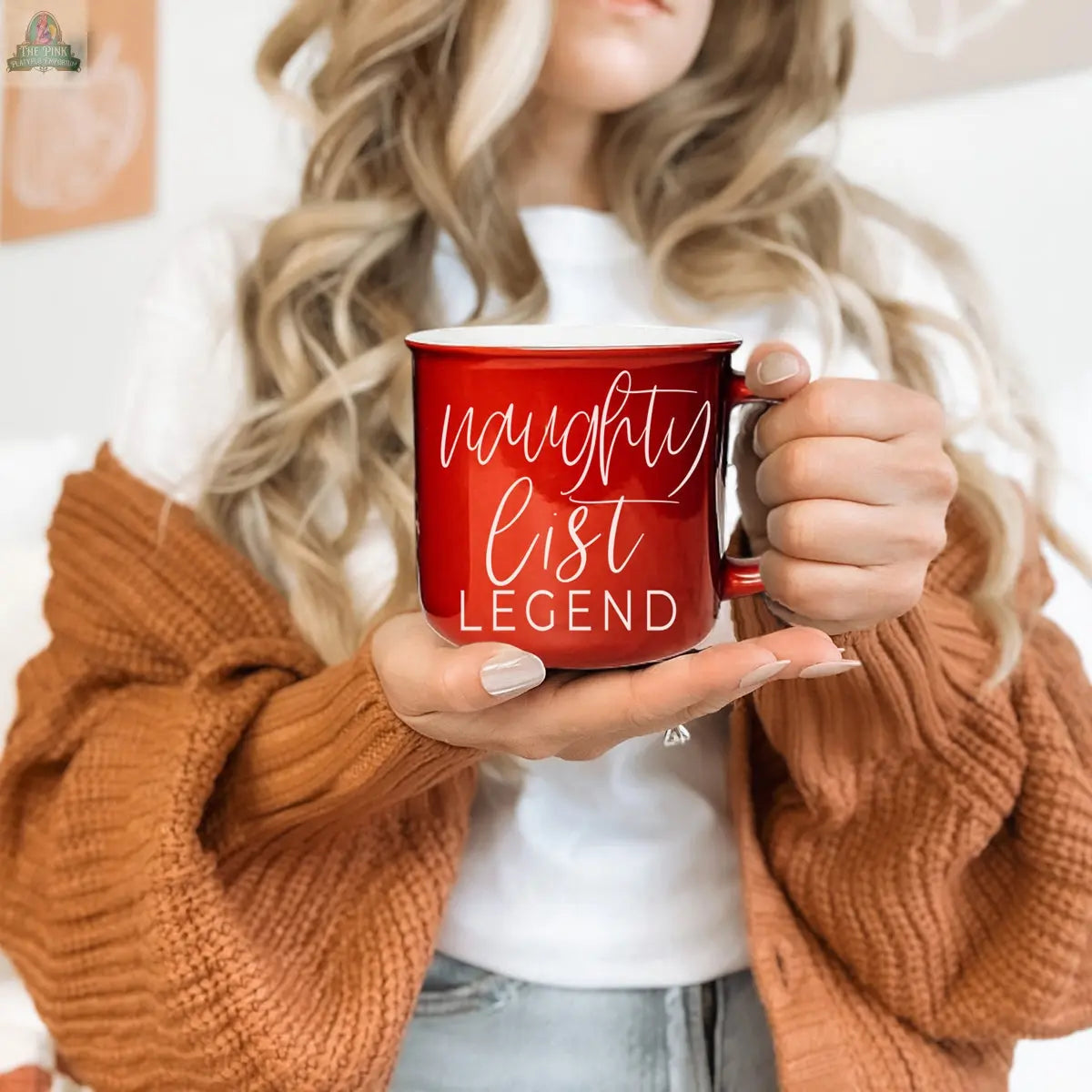 A woman with wavy blonde hair holds a festive Naughty Legend Mug. She wears a white shirt, an orange-brown knit cardigan, and light beige nail polish. The softly blurred background enhances the cozy holiday vibe.