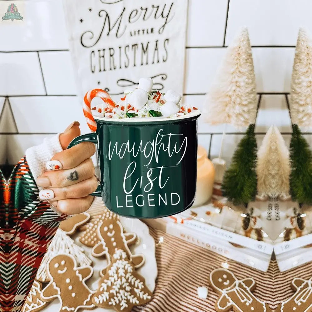 A hand with festive nails holds a Naughty Legend Mug filled with hot chocolate, marshmallows, and a candy cane. A decorated gingerbread cookie and a Merry Christmas sign add to the cozy holiday scene in the background.