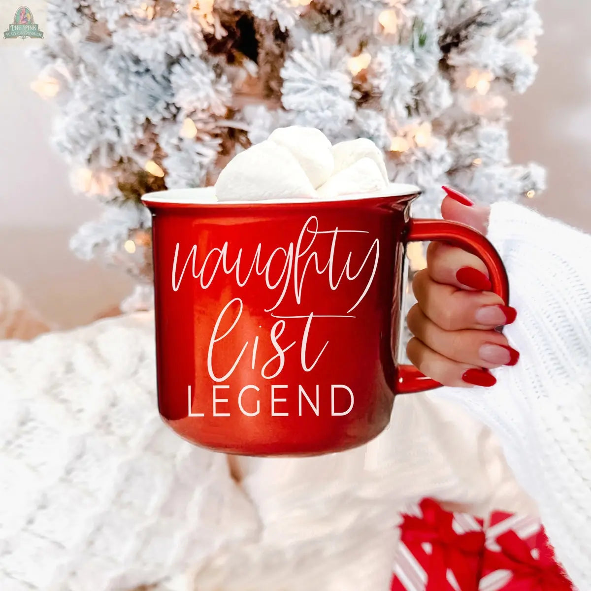 A hand with red-painted nails holds a Naughty Legend Mug filled with marshmallows in front of a white Christmas tree. The ceramic campfire mug reads "naughty list legend," adding festive charm to the bright holiday decor.