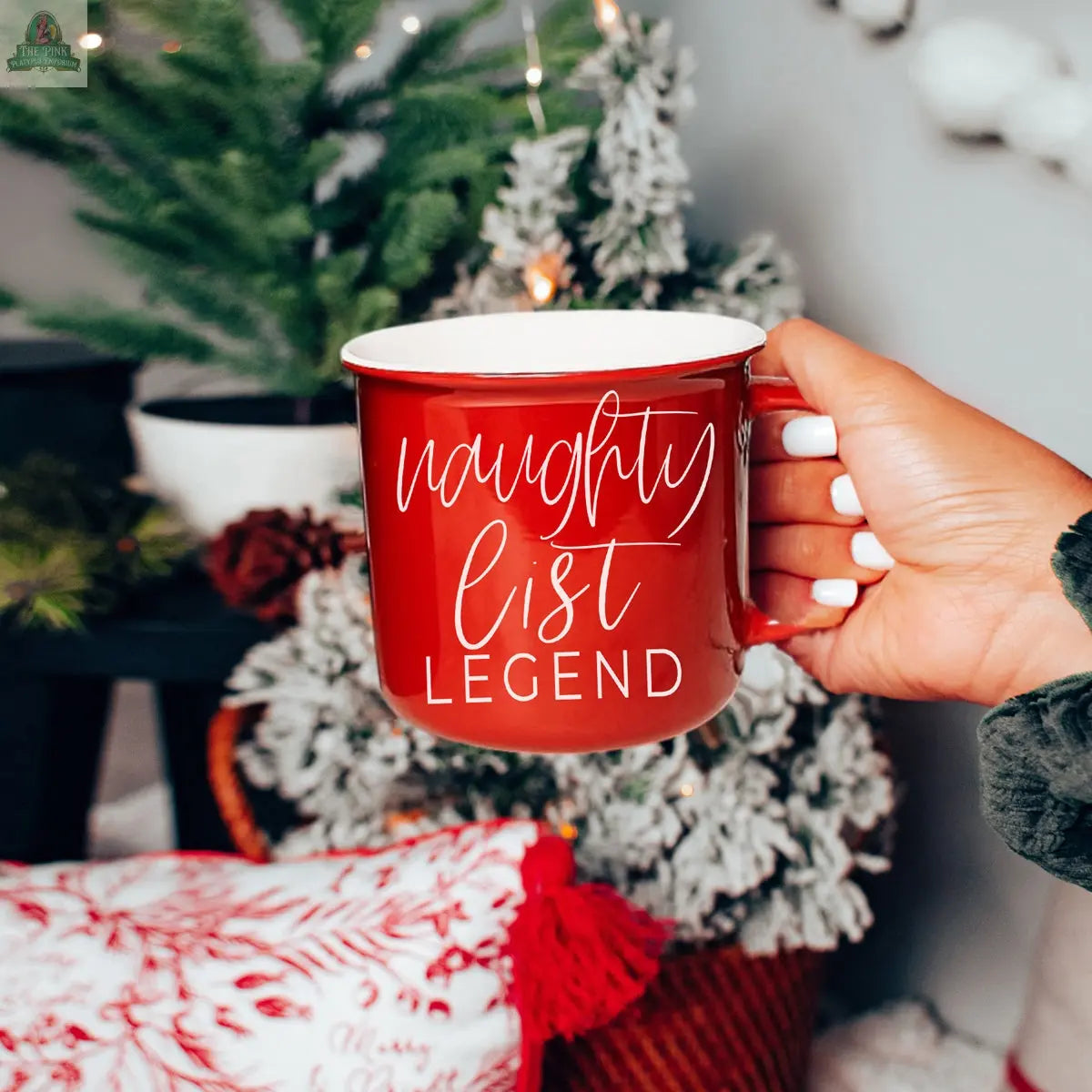 A hand with white-painted nails holds a red Naughty Legend Mug reading "naughty list LEGEND" in white script, with a festive Christmas tree and holiday décor in the background.