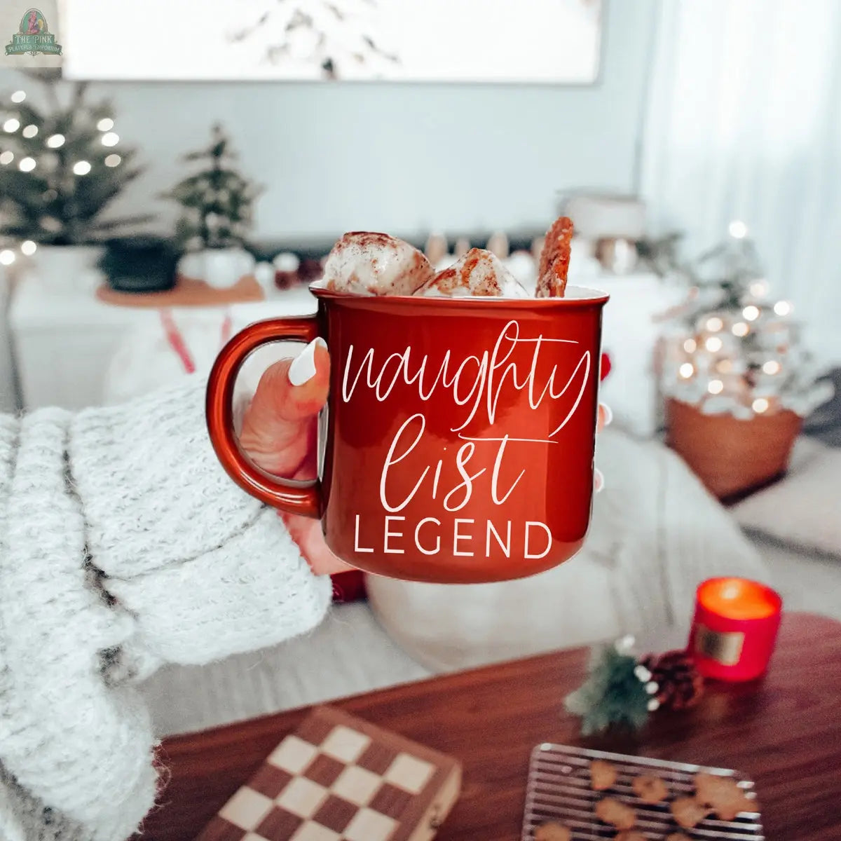 A hand in a cozy sweater holds a Naughty Legend Mug topped with whipped cream and a cinnamon stick. In the background, holiday decorations, a lit candle, a chessboard, and a small Christmas tree add festive cheer.