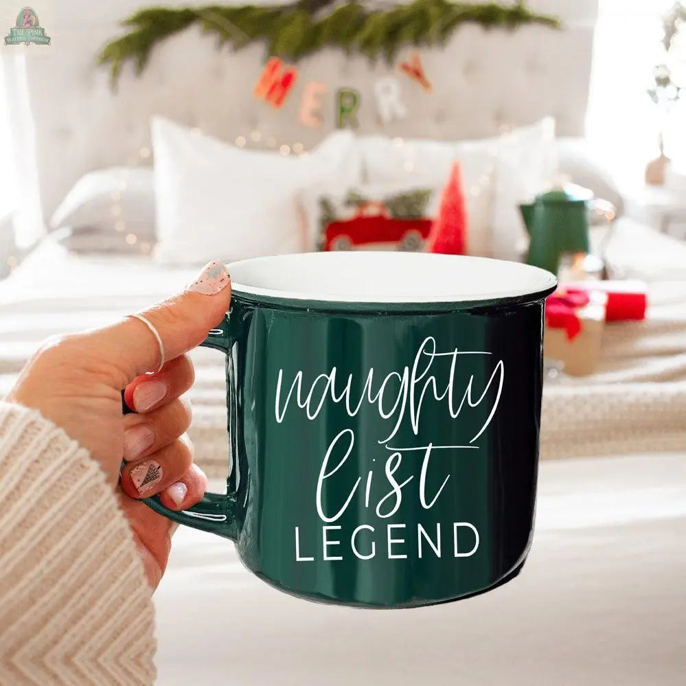 A hand holds the Naughty Legend Mug, a dark green cup with white "naughty list LEGEND" text, in front of a festive bed decorated with a MERRY banner and Christmas ornaments.