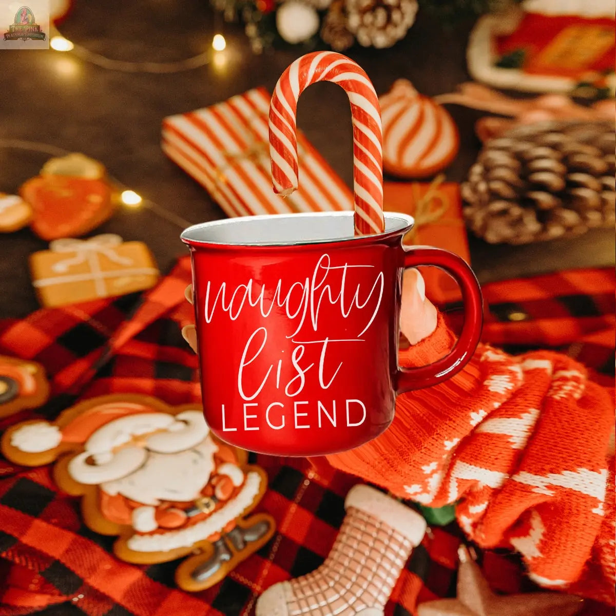 A person in a red and white striped glove holds a Naughty Legend Mug with a candy cane. Surrounded by festive decorations, cookies, and twinkling lights, this ceramic campfire mug is perfect for the holiday season.