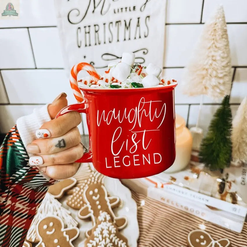 A hand with festive nail art holds a red Naughty Legend Mug filled with hot chocolate, marshmallows, and a candy cane. Gingerbread cookies and Christmas decorations are in the background.