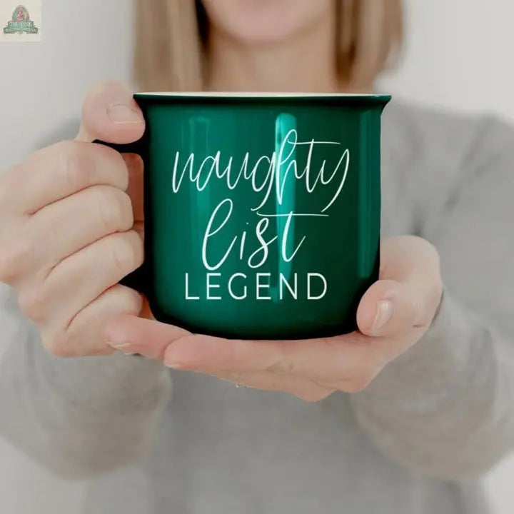 A person in a gray shirt holds the Naughty Legend Mug, a green cup with white text that reads “naughty list LEGEND.” The background is neutral and out of focus.