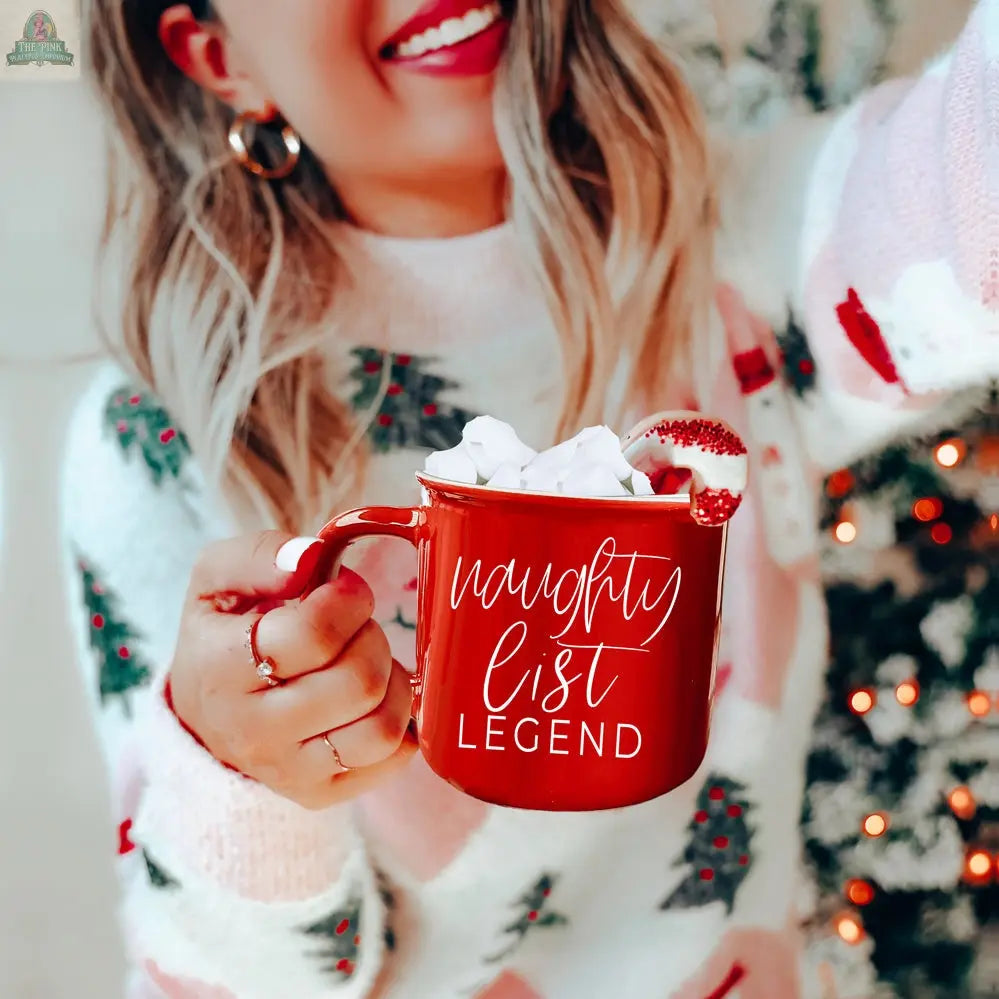 A smiling woman in a festive sweater holds a Naughty Legend Mug filled with marshmallows and topped with a candy cane. A blurred, decorated Christmas tree in the background adds a cozy holiday vibe.