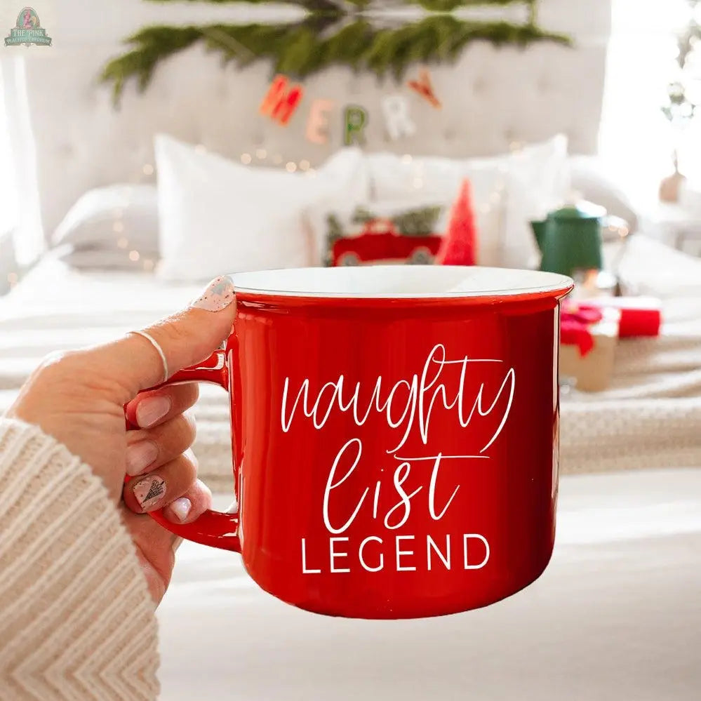A hand holds the Naughty Legend Mug, a red ceramic campfire mug with "naughty list LEGEND" in white script, in front of a cozy bed decorated for the holidays with wrapped gifts and a MERRY banner.