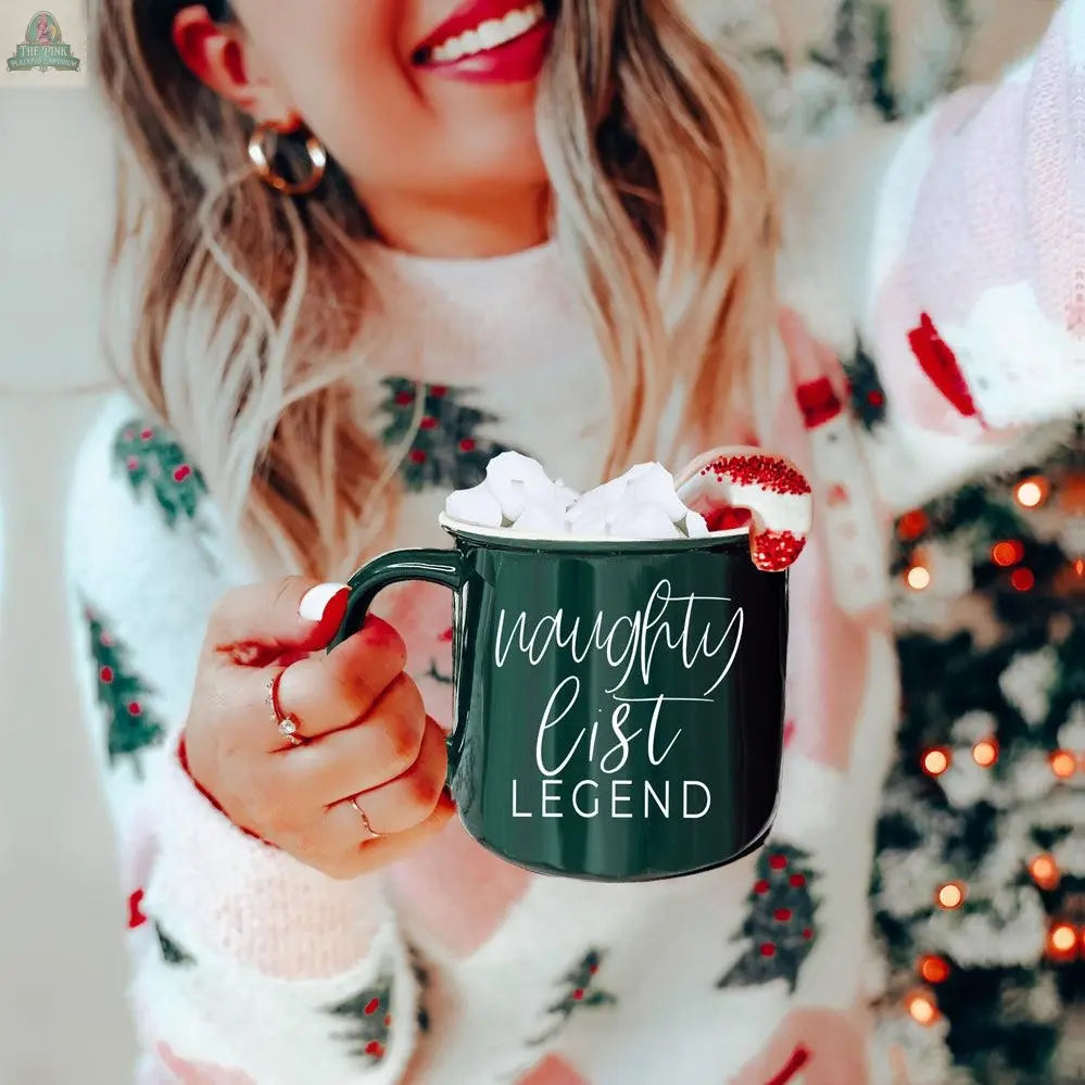A smiling woman in a festive sweater holds the Naughty Legend Mug, filled with marshmallows and a candy cane. The ceramic campfire mug reads "naughty list LEGEND," with a glowing Christmas tree in the background.