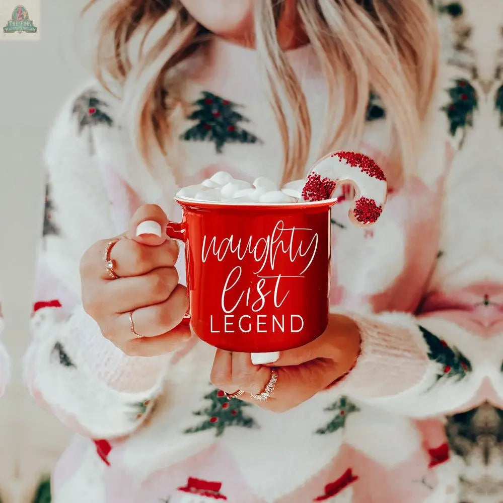A woman in a festive sweater holds a Naughty Legend Mug filled with marshmallows and topped with a donut coated in red and white sprinkles. This ceramic campfire mug features playful white text, making it the perfect holiday treat.