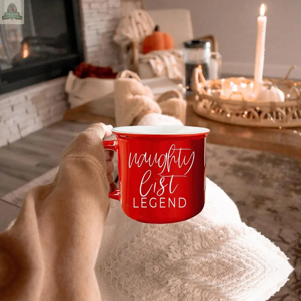 A person holds a red Naughty Legend Mug with white script, relaxing on a cozy blanket beside a lit candle and autumn decor in a warm, inviting living room.