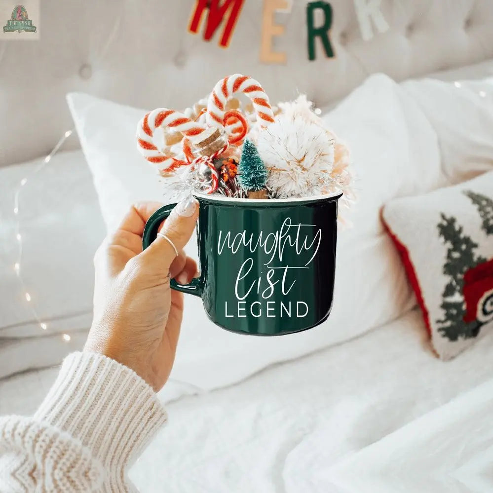 A hand holds the Naughty Legend Mug, a green ceramic campfire mug filled with Christmas treats and decor. In the background, festive bedding with holiday pillows and a MERRY banner enhance the holiday atmosphere.