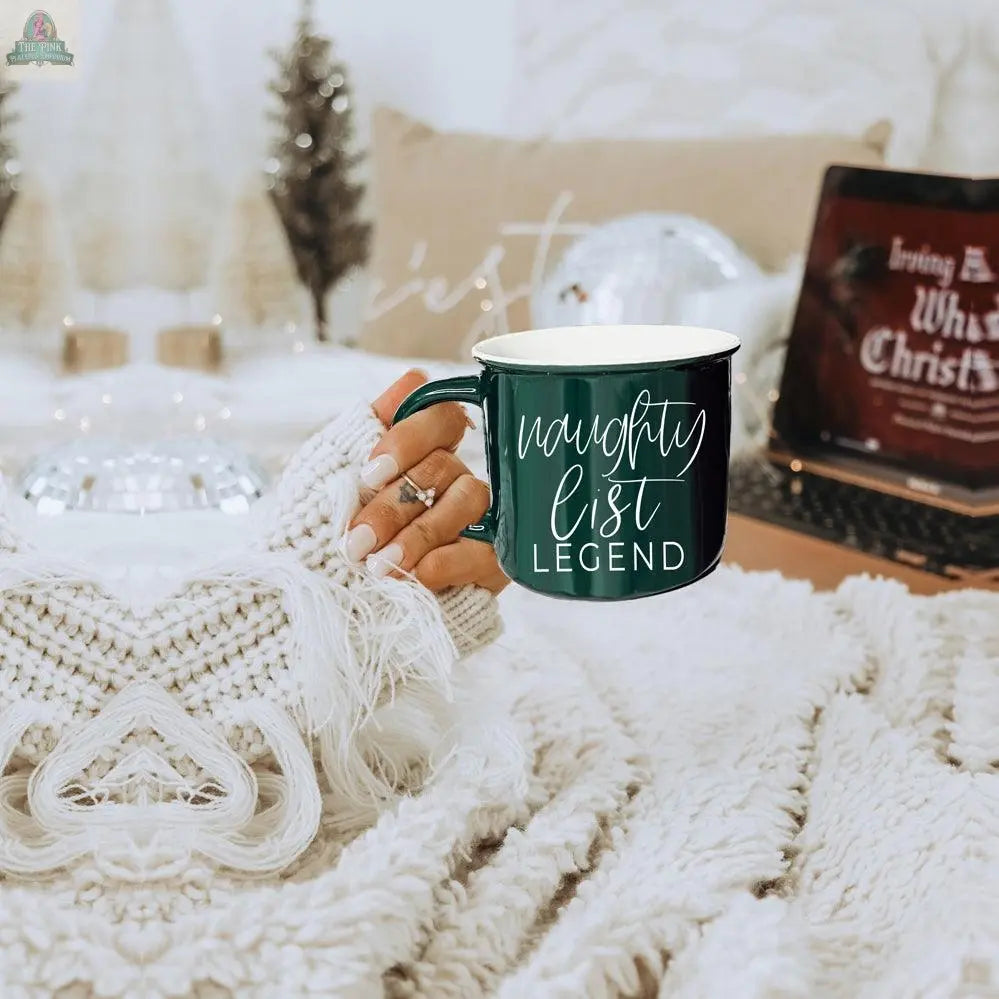 Wearing a cozy cream sweater, someone holds the Naughty Legend Mug—a festive green ceramic mug with “naughty list LEGEND”—while a laptop streams a Christmas movie amid charming holiday decor.