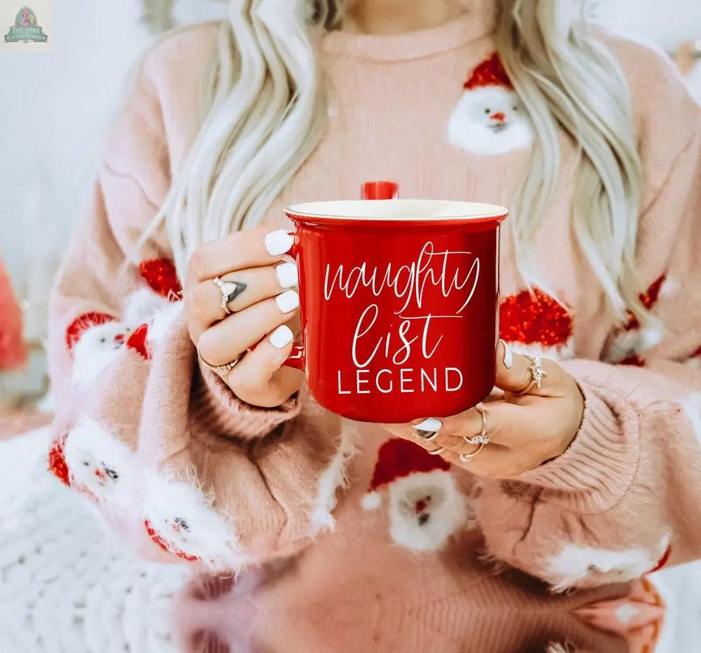 A woman with long blonde hair holds the Naughty Legend Mug, a red ceramic campfire mug. She's wearing a pink sweater with Santa Claus faces. Only her torso and hands are visible.