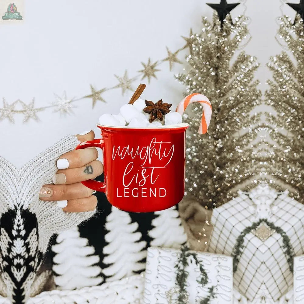 A hand in a cozy knit glove holds the Naughty Legend Mug filled with marshmallows, a candy cane, cinnamon stick, and star anise. Festive trees, gifts, and star decorations set the perfect holiday mood.