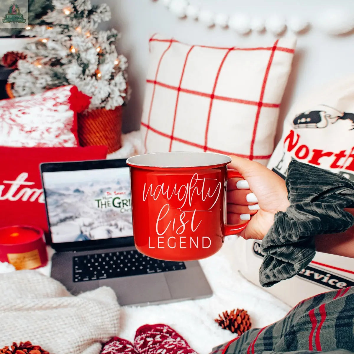 A hand holds a red Naughty Legend Mug in front of a laptop playing a holiday movie, with festive pillows, a candle, pinecones, and a decorated Christmas tree. This ceramic campfire mug adds charm to the cozy scene.