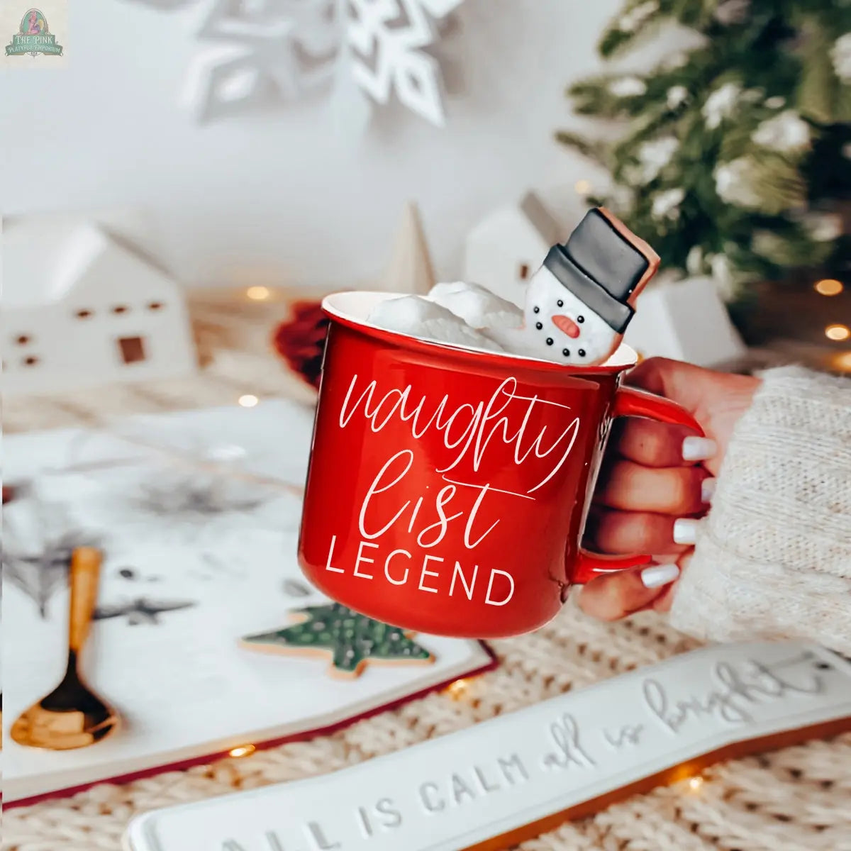 A hand holds a red Naughty Legend Mug filled with marshmallows and a snowman-shaped cookie. Holiday decorations, a tree, and cozy lights illuminate the festive background.