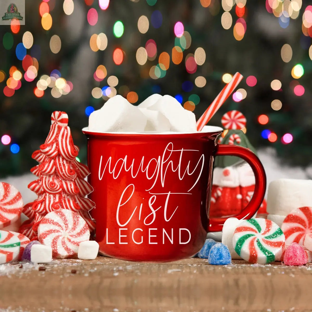 A red Naughty Legend Mug, ceramic campfire style, is filled with a candy cane and surrounded by colorful holiday candies and marshmallows. Blurred Christmas lights in the background create a festive, cozy atmosphere.